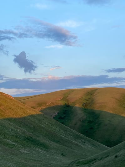 The image depicts a serene landscape of rolling green hills under a pale blue sky dotted with scattered clouds. The hills are covered in lush grass, with shadows cast across their slopes, suggesting either early morning or late afternoon light. In the foreground, the slopes are steep and textured, while the background reveals gentler, undulating contours. The sky is a gradient of soft blue, with wispy, elongated clouds and a singular, darker, amorphous cloud formation in the upper left quadrant. The scene is devoid of any man-made structures or human presence, emphasizing a natural and untouched environment.
