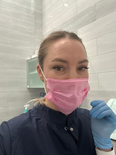 A person with light skin and blonde hair is wearing a pink surgical mask and dark blue medical scrubs. They are also wearing blue medical gloves on their right hand. The background consists of light grey vertical tiles and what appears to be a modern wall-mounted sink with a mirror. The setting appears to be a medical or dental office. There is no information to suggest the location is Kraskovo, Russia.