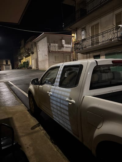 At night in Laureana di Borrello, Italy, a white vehicle is parked beside a curb on an asphalt street. Buildings with wrought-iron balconies line the street, lit by streetlights. Reflections on the wet pavement indicate recent rain. Another vehicle is partially visible on the right.
