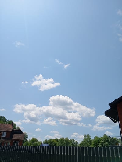 A clear blue sky with scattered white clouds dominates the upper portion of the image. A bright sunbeam angles down from the top left. In the lower part of the frame, a dark wooden fence runs horizontally across the foreground. Behind the fence, lush green trees form a dense treeline. To the left, a multi-story brick house with a red-tiled roof is visible, featuring several windows and scaffolding on one side. To the far right, the dark angled roofline and upper section of a modern brick building are visible. A small blue-tarped structure can be seen peeking out from behind the trees in the middle ground. The scene appears to be outdoors on a sunny day, likely during daytime, given the bright sunlight. The setting suggests a residential area with houses and natural greenery. No people are visible in the image, and there is no visible text. The overall impression is a bright and clear summer day in a suburban or rural setting.