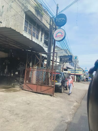 The scene is a street in Purwakarta, Indonesia, captured from a vehicle. The location is determined by the coordinates provided: latitude -6.543, longitude 107.441. On the left side of the street, there is a building with a sign that reads "Umami Coffee Shop". The building also has a large, red metal gate that is closed. On the opposite side of the street, a woman in a white hijab and a blue and white patterned dress is walking away from the camera. There is a white truck parked on the side of the street with "PURWAKARTA BURUT" written on it. Behind the truck is a sign for "PURWAKARTA DIESEL". On the street, there are electrical wires hanging overhead. There are also several other buildings in the background, including a building with the sign "Lively". The scene is captured on a bright, sunny day. The street is paved with concrete. The overall tone of the scene is mundane and ordinary.