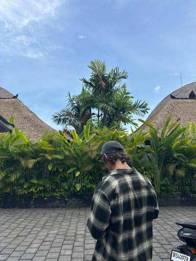 An outdoor shot in Tibubeneng, Indonesia, showcases a person wearing a green and black plaid shirt and a dark cap, standing next to a paved path. Lush green plants and a palm tree line the path. Above, a roof thatched with straw is visible against a blue sky with light clouds. A motorcycle is parked nearby, its license plate reading "DK5448FER".
