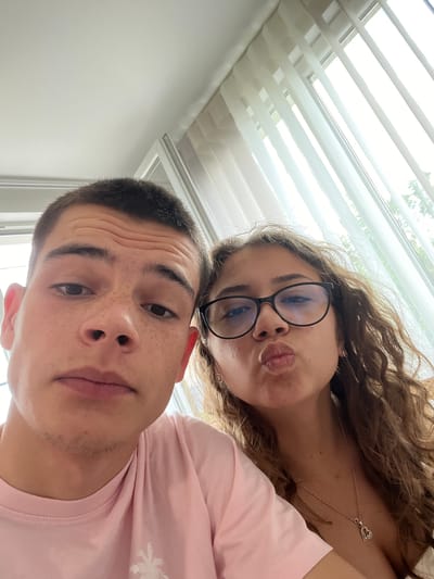 A selfie shows a young man and woman positioned close to each other. The man is in the foreground, and the woman is behind him, slightly angled. The man is wearing a pink t-shirt and has short dark hair. He has visible freckles. The woman is wearing glasses and is pursing her lips in a kissing expression. She has long, curly brown hair and is wearing a necklace with a heart pendant. In the background, there is a window with closed horizontal blinds, providing a source of light. The location is indoors. Karavelovo, Bulgaria, is not discernable from the image.