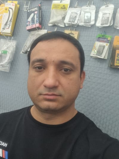 The image is a close-up, waist-up shot of a man with dark hair and a medium complexion, wearing a black t-shirt. He is standing in front of a wire grid wall with various products displayed on it. The products appear to be electronic accessories, such as phone chargers, cables, and batteries, some of which are packaged. The lighting suggests an indoor environment. The man's expression is neutral. The location is specified as Zafar, Uzbekistan, though there are no specific visual cues in the image to confirm this location. The man is the primary subject, and the background consists of retail display items.