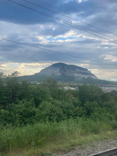 The image captures a view of Mount Zmeyka in the city of Zmeyka, Russia, under a dramatic sky filled with clouds. In the foreground, lush green trees and tall grass line the edge of a railway track. Beyond the trees, a town with houses is visible, nestled at the foot of the imposing mountain. The mountain itself is a prominent feature, with its slopes showing patches of lighter rock and vegetation. The sky above is a mix of dark storm clouds and areas where sunlight breaks through, creating a dynamic and somewhat moody atmosphere. Overhead, several power lines stretch across the frame, adding a man-made element to the natural landscape. The overall scene depicts a rural or semi-rural setting dominated by the natural grandeur of the mountain and the vast sky.