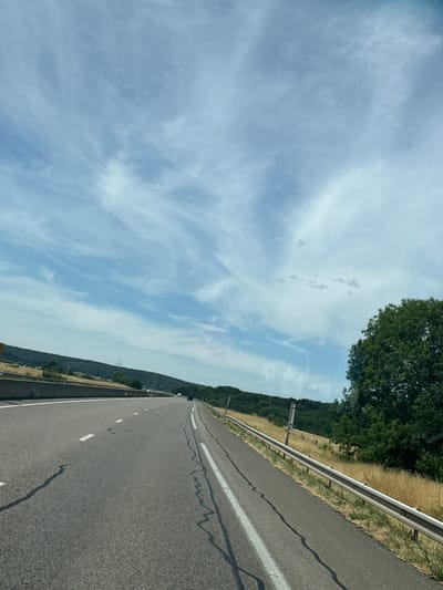 This is a forward-facing, eye-level, medium-angle shot of a paved highway in Manheulles, France. The highway is divided into lanes with white dashed lines. On the right side of the highway, there is a metal guardrail, and beyond that, dry grass and trees. A large green tree is prominent in the foreground on the right. In the distance, there are rolling hills covered with dark green trees. The sky is blue with wispy white clouds. There are no people visible in the image. The scene appears to be a rural roadside view.