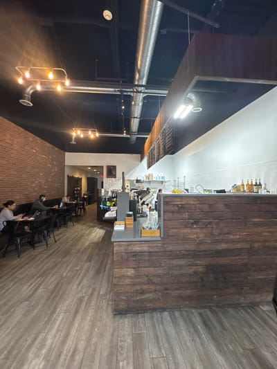 This image depicts the interior of a commercial establishment, likely a cafe or coffee shop, located in South Bend, United States.
The scene presents an industrial-chic aesthetic with a long, dark wood-paneled service counter on the right, featuring a light gray countertop and an espresso machine with associated equipment behind it. Above the counter, a menu board with white text on a black background is affixed beneath a dark wooden bulkhead. Behind the counter, a white subway tile backsplash extends along the wall, displaying various syrup bottles and other items. Overhead, the ceiling is painted black, exposing extensive silver-colored HVAC ductwork and a mix of industrial-style pendant lighting with exposed bulbs, alongside recessed and fluorescent fixtures.
To the left, a long, exposed red brick wall runs parallel to the counter. Along this wall, a dark cushioned booth seating arrangement is complemented by several small, square dark tables and matching dark metal chairs. Five individuals are visible in this seating area. Two individuals in the foreground, one wearing light gray clothing and another wearing green, are seated at separate tables, both actively engaged with open laptops. Further down the seating area, three additional individuals are partially visible, one also appearing to use a laptop.
The flooring throughout the establishment consists of light brown, wood-plank style laminate or vinyl. In the background, past the seating area and service counter, an open doorway leads into another dimly lit area, possibly a hallway or additional seating space. A single staff member is partially visible behind the counter in the distance, attending to the service area. No direct interactions between the seated customers are discernible; their primary actions involve individual use of electronic devices.