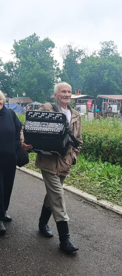 An elderly man with white hair and a weathered face, wearing a brown jacket, grey trousers, and black boots, is walking outdoors and carrying a black accordion. He is accompanied by a person on his left, whose face is not fully visible, wearing a dark top. They are on a paved path with greenery and trees in the background. Structures resembling stalls or small buildings are visible further back, suggesting an outdoor market or fair. The sky is overcast. The image is set in Старые Быги, Russia.