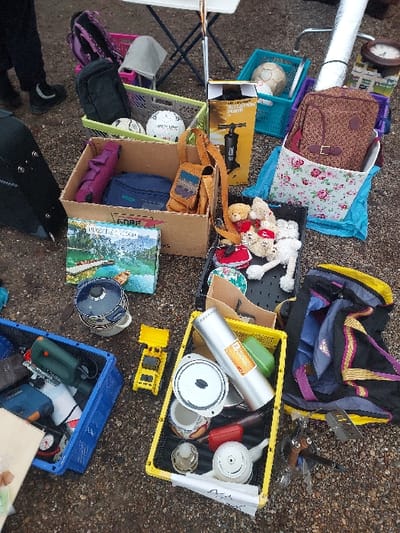 The scene depicts an outdoor flea market stall on a gravel surface in Eckbolsheim, France. In the upper left quadrant, the lower legs and feet of one individual are visible, clad in dark pants and dark shoes featuring a light-colored crescent moon emblem. A white folding table with metallic legs stands partially in the background.
Numerous items are displayed directly on the ground, primarily contained within various plastic crates (blue, green, yellow, black, pink) and cardboard boxes. Key objects observed include:
* **Containers:** Multiple crates and a floral-patterned fabric bin.
* **Sporting Goods:** Two white soccer balls (one branded "adidas"), a deflated soccer ball, and a boxed "INFLATION PUMP."
* **Bags/Packs:** A purple backpack, a black soft-sided bag, a brown leather-look shoulder bag with embroidered details, a brown speckled backpack, and a large dark duffel bag with purple and yellow accents.
* **Household/Miscellaneous:** A dark blue lidded pot, a white ceramic bowl, a white teapot, various other ceramic containers, red-handled kitchen utensils, a large silver cylinder (possibly a thermos), a brown wooden-framed wall clock, a silver rolled mat, and a white folded umbrella.
* **Tools:** A green and black electric jigsaw, identified as a Bosch brand tool.
* **Media/Toys:** A book or board game box titled "GORF" with a landscape image, a stack of optical discs, several small stuffed animals (bears and cat-like figures), and a small yellow toy excavator.
No explicit actions or interactions are depicted beyond the static arrangement of items for sale.