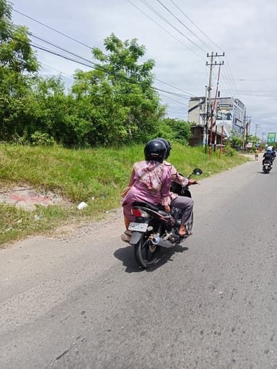 This is a daylight, eye-level, medium shot of two individuals riding a motorcycle on a paved road in Cotbadatunong, Indonesia. The rider and passenger both wear black helmets. The passenger is in front, wearing a light pink blouse with a floral pattern and grey pants. A light brown shoulder bag is slung over her left shoulder. The motorcycle appears to be a dark color. The road is bordered by a grassy embankment on the left and a line of trees. In the background, buildings and utility poles are visible, with a large banner on one of the buildings. There are other motorcycles in the distance on the road. The sky is cloudy.