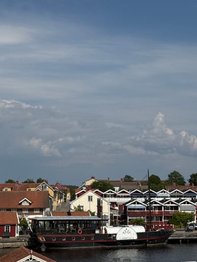 This is a medium-shot photograph of the harbor area of Askersund, Sweden, on a partly cloudy day. In the foreground, a dark-hulled boat with a covered upper deck and red trim is docked. The boat has a paddle wheel at its stern, and the name "E NORDEVALL II" is visible on it. Behind the boat, a row of red-roofed buildings with red and yellow facades line the waterfront. Further back, a street slopes uphill, with more buildings on either side, also featuring red roofs and colorful walls. Trees are visible interspersed among the buildings, and the sky above is a mix of blue and white clouds. The overall scene depicts a tranquil harbor town.