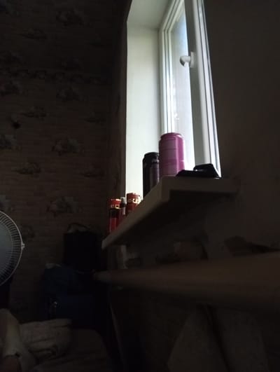 The image depicts an interior scene, likely a room, with a window on the right side. The overall lighting is dim, suggesting it could be nighttime or a poorly lit interior.
On a shelf in front of the window, several items are visible. From left to right, there is a dark-colored can, a pink can, and a dark object that might be a remote control. To the left of the shelf, partially obscured by shadow, are three red cylindrical objects with gold caps, possibly spray cans or bottles. Further left, a white fan with a wire cage is visible.
Behind the shelf and to the left, the wall is textured, resembling brick or stone with a pattern. A dark, rectangular object is also present in this area, possibly a piece of furniture or an appliance. In the lower left quadrant, a person's leg and foot are visible, covered by a light-colored blanket or clothing.
The scene does not provide explicit clues about the city of Yenakiieve, Ukraine. The description is based solely on the visual elements present in the photograph.