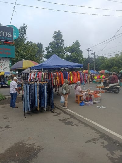 A street market in Purwokerto, Indonesia, located at coordinates -7.418 latitude and 109.251 longitude. Multiple stalls are set up with vendors selling a variety of goods, including clothing. There are three people visible in the frame: a man in a blue shirt and jeans standing near a clothes rack, a woman in a pink shirt and blue jeans standing near a rack of jeans, and a woman in a pink and white patterned shirt sitting at a table with a red tablecloth. The stalls are all shaded by canopies, and the ground is paved. There are power lines overhead. In the background, a motorcycle is parked near the edge of the scene, with a man in a blue shirt and jeans standing nearby. The sky is overcast and gray.