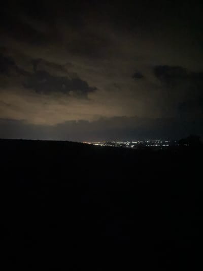 A night landscape photograph shows a dark foreground of rolling hills. In the distance, a city skyline is visible, illuminated by numerous lights. The sky above is dark with patches of clouds, some of which are illuminated from below by the city lights. The image was taken from a high vantage point looking down at the city of Sireți, Moldova.