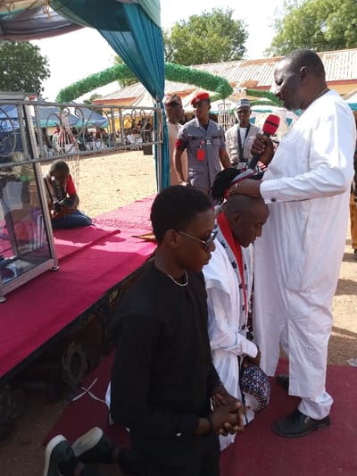 The image shows a scene outdoors in Zum, Nigeria, located at coordinates 10.116 latitude and 13.046 longitude. A man wearing a white robe and a microphone is speaking to a kneeling man. The kneeling man is wearing a traditional white robe with a colorful pattern, and he is holding a dark blue hat. To his left, a man is kneeling with his head bent and looking to the side. The man is wearing a black shirt, black pants, and sunglasses. A man in a black shirt is taking a picture with a camera. In the background, two men in red and white uniforms are standing. The event is taking place under a blue awning with green fabric hanging down the sides. The ground is covered in a red carpet. There is a metal box to the left of the scene. The image was taken on a bright, sunny day. A crowd of people are gathered around the event, and there are trees and buildings in the background.