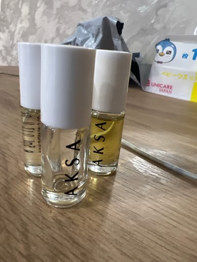 The image displays three small perfume bottles with white caps and clear glass bodies, arranged on a wooden surface. The bottles contain a yellowish liquid. The brand name "AKSA" is visible in black lettering on two of the bottles. The lighting suggests an indoor setting. To the left of the bottles, a crumpled grey plastic bag is partially visible, and behind that, a white package with a cartoon penguin and Japanese text including "UNICARE JAPAN" is seen. There are no people or any specific events depicted. The background is a wooden table with a subtle wood grain pattern. There are no immediate cues to suggest the time of day, weather, or specific emotions. The location context of Qoyankus, Kazakhstan, is provided but not visually evident in the image itself.