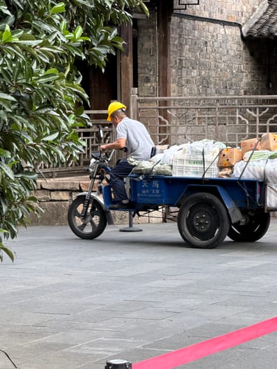 A male individual in a yellow hard hat, grey t-shirt, and dark trousers operates a blue, motorized cargo tricycle on a grey stone-tiled paved surface in Tongxiang, China. The tricycle, featuring a motorcycle-style front and a rear cargo bed, is heavily laden with white plastic crates, bundles wrapped in plastic or fabric, and cardboard boxes, secured by straps. The man is positioned with his back partially to the viewer, gripping the handlebars, and appears to be in motion. In the immediate foreground, a red barrier tape extends horizontally from a black stanchion on the right. Lush green foliage partially obscures the left side of the scene. The background reveals traditional Chinese architecture, including a dark grey stone building wall, wooden structural elements, and an ornate, dark lattice-patterned railing.