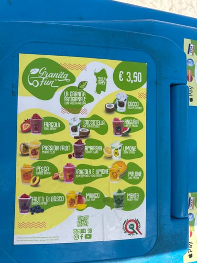 The image shows a bright yellow menu board advertising "Granita Fun" drinks, affixed to a textured blue plastic surface, likely a cooler or kiosk. The menu features various fruit flavors like strawberry, coconut, passion fruit, cherry, lemon, peach, strawberry and lemon, berry's family, mango, mint, and melon, each with a playful cartoon character in its cup. The price is listed as € 3.50. The text indicates the granita is artisanal and made with fresh fruit, and "Made in Italy." Payment icons for Visa and Mastercard are visible on the right side of the blue surface, along with the words "Pay" and an Apple Pay logo. A QR code and social media icons (Instagram, Facebook, YouTube) encourage customers to "Seguici Su" (Follow us). A seal at the bottom left indicates "Eccellenza Italiana 2021." The background is out of focus but appears to be outdoors, possibly a beach or tourist area, given the context of "Crabonaxa/Villasimius, Italy" which is a coastal area in Sardinia known for tourism. The image does not contain any people.
