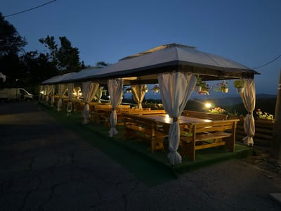 The image depicts an outdoor seating area at dusk, likely a restaurant or recreational space, located near Bogoslov, Bulgaria. The scene is dominated by a long, white-canopied gazebo structure, supported by poles and draped with light-colored curtains. Beneath the canopy, a series of wooden picnic tables and benches are arranged on a green carpeted surface. Hanging baskets with flowering plants are suspended from the gazebo's frame. The area is illuminated by artificial lights, casting a warm glow on the tables and benches. In the background, rolling hills are visible against a darkening blue sky, and trees line the edge of the property. To the left, a white van is parked. The foreground shows a cracked asphalt surface. There are no people visible in the image.