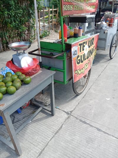 The scene takes place on a concrete sidewalk in the city of Kendal, Indonesia, located at coordinates -6.931 latitude and 110.204 longitude. A street food vendor has set up shop with a green and silver cart. The cart is equipped with a scale and a large sign advertising "TELUR GULUNG", which appears to be a local food item. There are several avocados on a gray table in front of the cart. Bottles of condiments are lined up behind the sign on the cart. The vendor is not visible in the image. The sidewalk is cracked and weathered.