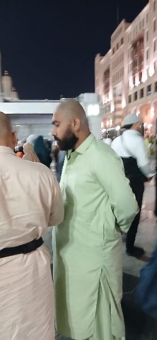 The image captures a nighttime street scene in Medina, Saudi Arabia, specifically in the vicinity of the Prophet's Mosque (Al-Masjid an-Nabawi), identifiable by the distinctive illuminated buildings and minaret-like structure in the background.

In the midground, a male individual is prominently featured in profile, facing right. He possesses a shaved head and a full, dark beard, and is dressed in a light green, collared, long-sleeved tunic (qamis or thobe) that extends past his hips. To his immediate left, partially visible from the back, is another male individual with a bald head, wearing a light-colored, possibly off-white, long garment fastened with a black belt. In the blurred background to the right, a person wearing a white top and dark lower garment with a light head covering is visible amongst other indistinct figures, some in traditional dress, suggesting a public, populated area.

The environment is illuminated by artificial light sources. The background showcases a large, multi-story building with numerous glowing windows, indicative of modern architecture commonly found around significant religious sites. On the far left, a white architectural element resembling a minaret, topped with a golden dome, stands against a dark night sky. The ground visible at the bottom of the frame is paved.