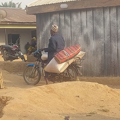 In Gudi, Nigeria, a man is depicted seated on a blue motorcycle on an unpaved dirt path. He wears a dark blue long-sleeved garment and a patterned, light-colored cap. His bare feet are visible near the ground. The motorcycle is heavily laden with cargo, including two large, light-colored sacks secured to the rear seat and rack, topped by a red and patterned cylindrical object.
In the background, to the left, a single-story building with a light-colored facade and a brown corrugated metal roof is visible. A red motorcycle is parked on a raised dirt embankment in front of this building. Two individuals are seated on the embankment near the building's open doorway; one is clearly visible wearing green patterned trousers and a yellow head covering. A satellite dish is mounted on the wall of this building.
To the right, another single-story building features a grey, textured, possibly corrugated metal exterior wall. A white oval-shaped light fixture is mounted on its exterior below the roofline. The ground throughout the scene is composed of dry earth with visible mounds, ruts, and sparse vegetation, indicating an unpaved surface.