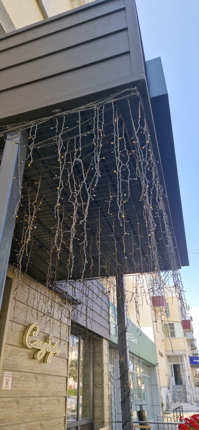 The image shows the exterior of a building in Miass, Russia. The building has a wooden facade and is decorated with strings of small lights, resembling icicles. A sign with the inscription "Сладости" (Sweets) is visible on the facade. Several shops and apartment buildings are visible in the background. The sky is clear and blue. The image appears to be taken during the day.