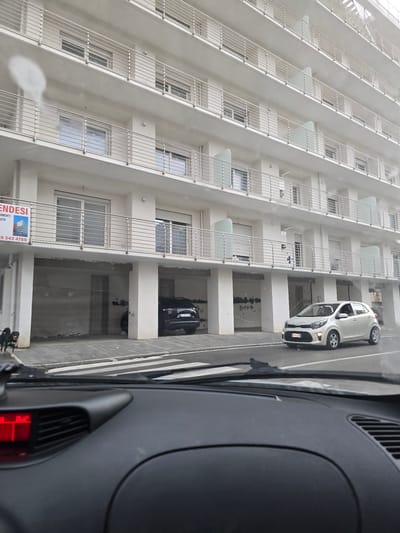 The image shows a modern, white multi-story building with numerous balconies and windows, likely an apartment building. In front of the building, a street with a crosswalk is visible. Two cars are present: a black SUV parked in a covered area beneath the building, and a small white car driving on the street. A dog is walking on the sidewalk to the left of the building. A "VENDESI" (For Sale) sign is attached to the building on the left side. The scene appears to be in a city, with the architecture suggesting a European location. The presence of the "VENDESI" sign and the overall urban setting are consistent with a location in Salerno, Italy.