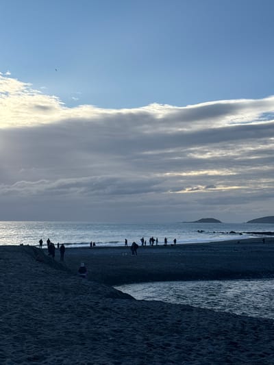 The image depicts a coastal scene at Seaton, United Kingdom. The sky occupies the upper two-thirds, featuring a clear blue expanse in the top portion, below which lies a horizontal layer of bright, white-edged clouds, transitioning into a dense stratum of grey clouds across the mid-sky. Sunlight penetrates the clouds from the left, creating a luminous area. A single dark bird is visible flying in the clear blue sky.
The sea, appearing dark blue-grey, exhibits gentle waves near the shoreline. Two distinct, dark landmasses, possibly islands or headlands, are visible on the horizon to the right.
The foreground consists of a dark-toned beach, likely composed of shingle or coarse sand. A prominent, dark sandy bank or ridge is present in the lower left. A narrow river or stream flows from the right foreground, curving left to meet the sea, forming a discernible channel of dark water.
Approximately 25-30 individuals are observed on the beach and at the water's edge, primarily in silhouette. They are distributed across the mid-ground, mostly standing or walking. Several individuals are accompanied by dogs. One smaller individual, consistent with a child, wearing a light-colored hat and jacket, is positioned low on the sandy bank in the near foreground. The overall lighting suggests late afternoon or early morning due to the low sun angle causing backlighting and reflections on the water.