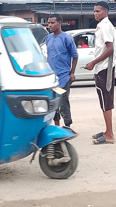 The image depicts a scene on a street in Warri, Nigeria. A blue and white auto rickshaw, also known as a "keke," is positioned on the left side of the frame. Two young men are standing near the auto rickshaw, seemingly engaged in conversation. One man is wearing a blue, short-sleeved shirt and dark pants, while the other is wearing a white, short-sleeved shirt, dark shorts with white trim, and sandals. A silver vehicle is visible in the background, with a person inside. The environment appears to be a typical urban setting in Nigeria, with a building visible behind the vehicles. The ground surface is concrete.