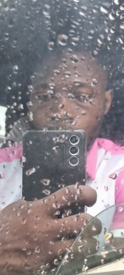 A selfie is taken through a rain-streaked window, presumably from within a vehicle. The subject, a person with dark, curly hair and medium-toned skin, holds a black smartphone in their right hand, using it to capture the reflection in the window. The person is wearing a pink and white collared shirt. The background is blurred due to the rain and the reflection, but the impression is of an outdoor, potentially urban environment. The context of Idimu, Nigeria, suggests this scene may be a common occurrence during periods of rainfall in that region.