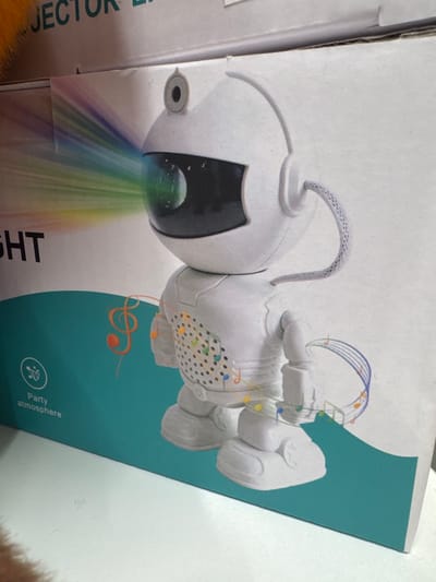 The image displays the packaging of a projector light product, likely a toy or decorative item. The dominant subject is a stylized, white robot figure depicted in a cartoonish manner. The robot has a round head with a dark, glowing lens from which a vibrant rainbow of light streams downwards. Its body is adorned with musical notes and colorful dots, suggesting it might project lights and play music. The robot is positioned on a teal-colored background which transitions to white on the right side of the package.

The packaging itself is primarily white, with sections of the teal design. Visible text on the packaging includes "JECTOR L" in large, dark letters on the left side, partially obscured by the edge of the box and a glimpse of orange fur. On the teal section, near the bottom right, smaller white text reads "Party atmosphere" next to a circular icon featuring a music note and sound waves. The overall impression of the product's design is playful and aimed at creating a festive or stimulating visual experience.

There are no people visible in the image. The setting appears to be indoors, possibly a retail environment or a home, given the lighting and the nature of the product. The image is a photograph of the product packaging, likely taken to showcase the item. No specific time of day, weather, or strong emotional cues are present beyond the cheerful and fun aesthetic conveyed by the product's design. The location context provided is Keila, Estonia, but there are no visual cues within the image itself to confirm this specific location.