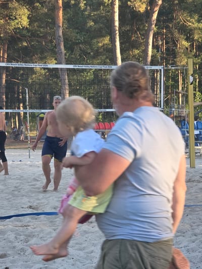 A man is holding a small child on a sandy beach, possibly a volleyball court, with a net and trees in the background. The man is wearing a light blue t-shirt and olive green shorts, with his back to the camera. The child is wearing a light blue shirt and a yellow skirt, and is facing away from the camera as well. The child's legs are visible, with one leg extended. A volleyball net is in the background, with text on it that reads "CEV Beach Volley Nations Cup Final 2024". There are trees behind the net, and some spectators in the background. The location is identified as Daugavpils, Latvia.