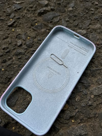 An overhead shot shows the interior of a light blue, soft-touch phone case with a faint iridescent sheen on its edges, resting on a dark, rough, and uneven surface. The phone case is positioned diagonally, with the top-left corner closest to the viewer. The interior is lined with a greyish-blue material and features embossed text and circular patterns. A small, silver SIM card ejector tool lies horizontally across the center of the case, resting on the textured interior.
Visible text on the interior of the case reads: "YOUR PHONE. YOUR PRIDE." and "LIQUID SILICONE + SUEDE ULTRAPRO". Another embossed line reads "LIQUID SILICONE" within a circular outline. The outer edge of the phone case has a subtle gradient of pink, purple, and blue.
The background is a dark, coarse ground composed of small, irregularly shaped stones and dirt, suggesting an outdoor environment. There are small specks of yellow material scattered on the ground, particularly in the lower right portion of the image. The lighting appears to be natural daylight, casting no strong shadows, indicating it could be daytime, possibly overcast. The overall scene is static, with no people or discernible activity, focusing solely on the object and its immediate surroundings. The location context of Reuleut Timu, Indonesia, is noted but not visually evident from the image itself.