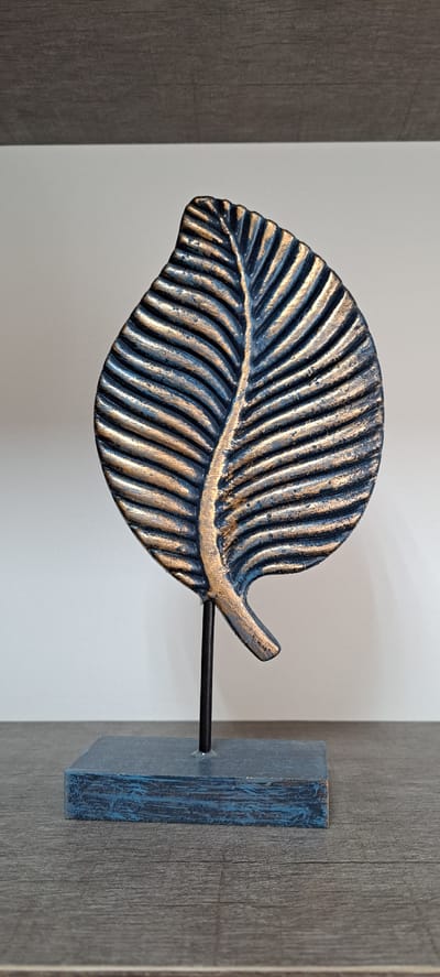 A dark blue and gold leaf sculpture is displayed on a dark gray surface in front of a white wall. The sculpture is a stylized representation of a leaf with prominent veins, rendered in textured dark blue with accents of metallic gold. It is mounted on a thin black metal rod, which is attached to a dark blue rectangular base. The base rests on a textured surface that appears to be a wooden shelf. The background wall is plain white, and above the white wall, a dark horizontal surface, possibly a shelf or cabinet, is visible. The lighting suggests a studio or indoor display environment, with no visible signs of the city of Yessentuki, Russia.