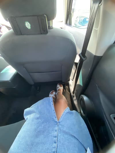 The image shows the interior of a vehicle, likely a car, viewed from the perspective of someone sitting in the back seat. In the foreground, a person's legs are visible, wearing a light blue denim skirt and white sandals. The person's feet are on the floor of the car. The back of the front passenger seat is in the center of the image, with a headrest. A green logo is visible on the headrest. To the right of the seat, a black seatbelt is buckled. The interior of the car appears to be a typical car interior with dark gray seats and a black floor mat. The environment outside the car is suggested by the bright light filtering through the windows, indicating a sunny day. The specific location of Yuldybayevo, Russia is not visually discernible from the image itself.