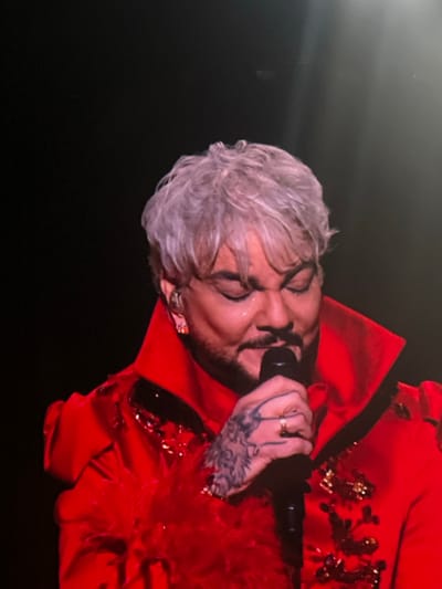 A male subject with platinum blonde or silver hair, a beard, and a mustache is depicted on a stage in Novoivanovskoe, Russia. He is dressed in a bright red collared jacket featuring red sequins and red feather cuffs, worn over a red undergarment. An in-ear monitor and an earring are visible in his left ear. His right hand grips a black microphone, displaying a gold-toned ring on the ring finger. Dark, intricate tattoos are visible on the back of his left hand. His eyes are closed or heavily squinted, and moisture is present on both cheeks. The background is dark, suggesting a performance setting, with a faint vertical light beam visible on the right.