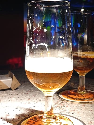 A close-up, low-angle shot captures two glasses of amber-colored liquid, likely beer, on a speckled countertop, possibly in a bar or restaurant in Hergiswil (NW), Switzerland. The primary glass, in the foreground, is a stemmed tulip glass filled to about two-thirds capacity with a frothy head. The liquid shows reflections of colorful lights in the background, which are out of focus. Small bubbles cling to the inside of the glass. To the right, a second, partially visible stemmed glass contains a darker liquid, with a slice of what appears to be lemon floating on the surface. Both glasses sit on circular coasters with orange and black branding. In the lower-left corner, a small, unfolded cardboard box is visible. The background is dark with blurred, colorful lights, suggesting an indoor setting with ambient lighting. The lighting casts a shadow from the foreground glass onto the countertop.