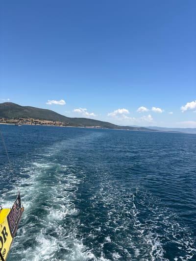The image presents a serene coastal landscape as seen from a moving boat under a clear, bright sky. The scene is dominated by a vast expanse of deep blue sea and a distant, undulating coastline.

In the upper part of the frame, a brilliant blue sky stretches across, adorned with scattered, fluffy white clouds that suggest fair weather conditions. The horizon line runs horizontally across the mid-ground, separating the sky and distant land from the water.

Along the horizon, a long stretch of land forms the coastline, featuring gentle hills and mountains. These landforms are covered in varied vegetation, appearing green and somewhat dry in parts, and fading into a hazy, lighter tone in the furthest distance.

The lower half of the image is filled with the dark blue waters of the sea. The surface of the water shows ripples and waves, with sunlight glinting off it. Towards the bottom right of the frame, a prominent white wake of churned water and foam trails behind the vessel, indicating its movement.

In the bottom right corner, a section of the boat from which the photo was taken is visible. It features a bright yellow object, possibly a fender or a part of the boat's exterior, with partially legible text and graphics. Visible letters include "DR" and "OR", along with numbers such as "68" or "89", and a QR code. A thin rope or cable extends from this part of the boat.

The time of day is clearly daylight, likely morning or early afternoon, given the intensity of the light and the clear sky. The overall impression is one of peaceful travel and scenic beauty, consistent with a coastal journey off Menderes, Turkey.