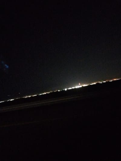 The image captures a nighttime view of the city of Jibek Joly, Kazakhstan, from a distance. The foreground is dominated by darkness, with the exception of a faint, dark, sloped surface that could be the ground or a road. In the background, a distant cityscape is visible, characterized by a horizontal line of illuminated lights. These lights are a mix of white, yellow, and orange, suggesting a concentration of buildings and urban infrastructure. The sky above is predominantly black, with a subtle blueish tint on the left side, possibly due to artificial light or atmospheric conditions. The overall impression is one of a vast, dark landscape punctuated by the glow of a city at night.