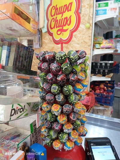 The image depicts a close-up view of a display of Chupa Chups lollipops in a store. The lollipops are arranged on a stand, with many individual wrappers visible. The wrappers feature various designs and colors, including some with "Black Edition" branding. In the background, shelves stocked with other products, including boxes of Dirol gum and what appear to be other confectioneries, are visible. There is also a blue crate filled with peaches and a payment terminal in the foreground. The setting appears to be a retail environment, possibly a small shop or a kiosk. The image was taken in Nyurba, Russia, as indicated by the text overlay "REDMI 13C | ИВАНОВ". The time and date displayed on the payment terminal are 04.07.2026 14:01.
