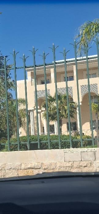 This image depicts a multi-story building with a light-colored, possibly beige or tan, facade situated in Berrechid, Morocco, under a clear blue sky. The building exhibits architectural elements typical of Moroccan design, including several prominent arched openings on the ground floor supported by columns. Between these arches and on the upper levels, there is decorative tilework featuring blue and white geometric patterns. The upper floor includes recessed windows or balconies.
In the foreground, a dark green metal fence with pointed, ornate finials runs horizontally, partially obscuring the view of the building. Behind this fence, there is a manicured strip of dense green bushes. Below the fence and bushes, a low retaining wall is visible, constructed from light-colored rough-cut stone blocks at the base, topped with a lighter concrete ledge that shows some white markings. Several palm trees and other green trees are positioned behind the fence and in front of the building, with one prominent palm tree centered between the building's arched entrances. No individuals are visible in the scene. A dark, indistinct surface occupies the bottom portion of the image, suggesting the photograph was taken from within a vehicle.