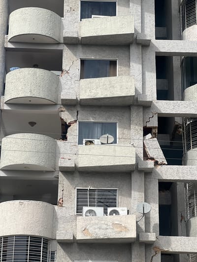 The image shows the facade of a concrete building with a textured, off-white finish. The architecture features repeating vertical elements that create a series of balconies or recessed openings on each floor. On the upper floors, some of these openings reveal darker interior spaces, and in some areas, there is visible structural damage, with concrete broken and exposing brickwork. Several windows are visible on the lower floors; some are dark, suggesting they are tinted or covered, while others show interior details like air conditioning units and grates. A satellite dish is mounted on one of the upper sections. Thin wires, likely for utilities or communication, are strung vertically across parts of the facade. The lower level of the building has rounded, projecting elements, possibly balconies. The lighting suggests it is daytime, with strong shadows indicating the sun's position. The overall impression is one of urban architecture showing signs of age and disrepair. The location is Maracay, Venezuela.