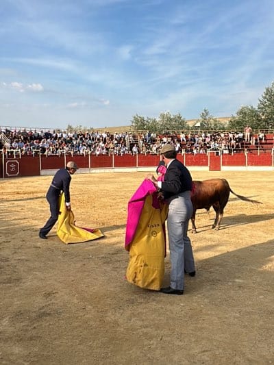 The image depicts a bullfight in an arena, likely in San José del Valle, Spain, given the cultural context. Two matadors dressed in traditional attire, one bending over with a yellow cape and the other holding a yellow and pink cape, are facing a bull. The arena is filled with spectators in the background. The setting includes a dirt ground and a red barrier surrounding the fighting area.