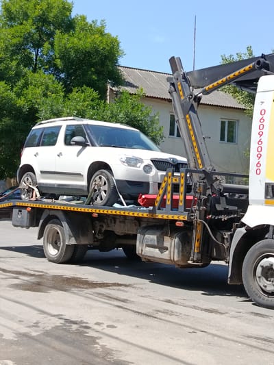 A white Skoda Yeti is being loaded onto a tow truck in the city of Bălți, Moldova. The tow truck is yellow and white and has a crane arm extended over the car. The car is secured to the tow truck with straps. The tow truck is parked on a paved road, with trees and a building in the background. The sky is blue and clear. The scene appears to be taking place during the daytime.