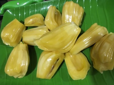 The image shows a pile of ripe jackfruit segments arranged on a bright green, textured platter. The jackfruit is a vibrant yellow, with a smooth, slightly glossy surface, indicating its freshness. Some segments have a lighter, almost creamy yellow hue, while others are a deeper yellow. The irregular shapes of the segments, with their characteristic ribbed texture, are clearly visible. A portion of a green knife or tool with a metallic handle is visible in the top left corner, suggesting preparation. The platter itself has a wavy pattern, and the overall lighting suggests an indoor setting, possibly a kitchen or dining area. There are no people visible in the image. The focus is entirely on the jackfruit and the presentation. The image likely captures a moment of food preparation or display, highlighting the appealing appearance of the fruit. Given the location context of Tingkeummanyang, Indonesia, this imagery is consistent with the availability and consumption of jackfruit in tropical regions.