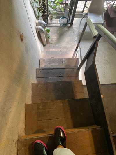 The image shows a person standing at the bottom of a wooden staircase in a building in Kecamatan Piyungan, Indonesia, at the coordinates -7.827 latitude and 110.482 longitude. The staircase is made of weathered wood with visible wear and tear. The person is wearing black shoes with pink accents, and the top of their white pants is visible. The staircase is positioned against a concrete wall with a portion of a metal railing visible to the right. There are also metal pipes and a potted plant visible to the left side of the staircase. The background shows a metal gate, suggesting that the staircase leads to a higher floor or level. The overall scene is a simple, everyday image of a person standing on a staircase inside a building.