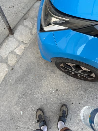 The image shows a first-person perspective of a person standing on a concrete sidewalk next to a blue car. The car is a compact electric vehicle, likely a Dacia Spring or similar, with its headlight visible. The front passenger wheel and part of the body are in the frame. The sidewalk is paved with grey concrete and has some uneven stone slabs near the curb. A metal pole, possibly a street sign or barrier, is visible on the left. The person is wearing cycling shoes and white socks. In the foreground, a plastic water bottle is held by an unseen hand. The scene appears to be an urban environment, and given the context, it's possible this is a street in La Llacuna, Spain. There is a discarded cigarette butt on the ground near the metal pole. The lighting suggests daytime.