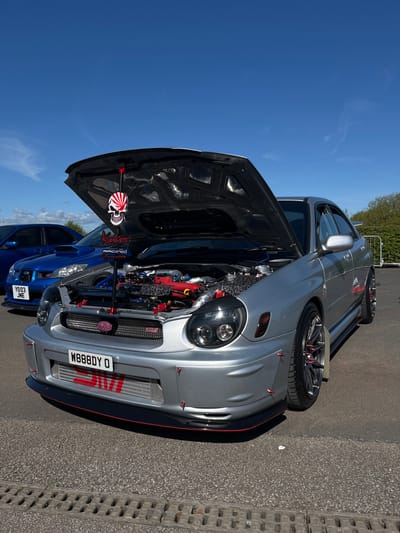 The image shows two Subaru Impreza WRX cars with their hoods open, parked on an asphalt surface in Silverstone, United Kingdom. The car in the foreground is silver, with a visible intercooler featuring red detailing and a license plate reading "W888DY O." The hood is held open by a prop rod, adorned with a red and white skull emblem and the word "Kamikaze" in vertical lettering. The engine bay displays various components and hoses in red and blue. Behind the silver car is a blue Subaru Impreza WRX, also with its hood open. The sky is blue with few clouds.