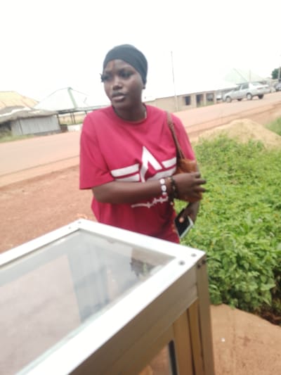 A young Black woman wearing a red t-shirt with a white abstract design and a black headscarf is standing outdoors. She has dark skin, wears false eyelashes, and has bracelets on her left wrist. She is holding a brown handbag on her left shoulder and a dark-colored smartphone in her right hand. In front of her is a white and aluminum display case with a glass top. The background shows a dirt road, some buildings with corrugated roofs, green vegetation, and a silver sedan parked on the road. The scene appears to be in Agwala, Nigeria.